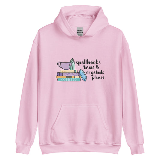 A pink hooded sweatshirt featuring an illustration of a stack of spellbooks with a teacup and a crystal resting on top. Another crystal rests on the side of the stack, and text next to the illustration reads "Spellbooks, teas & crystals please"
