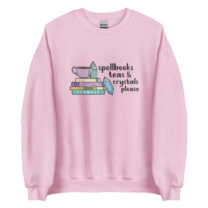 A pink crewneck sweatshirt featuring an illustration of a stack of spellbooks with a teacup and a crystal resting on top. Another crystal rests on the side of the stack, and text next to the illustration reads "Spellbooks, teas & crystals please"