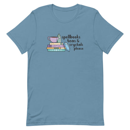 A blue-gray t-shirt featuring an illustration of a stack of spellbooks with a teacup resting on top. A few crystals are scattered around the books. Text alongside the illustration reads "Spellbooks, teas & crystals please"