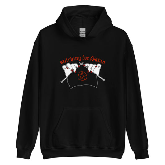 A black hooded sweatshirt featuring an illustration of pale white hands with red fingernails holding knitting needles. Fabric on the needles features a red pentagram. Text above the hands reads "stitching for Satan" in a gothc red font.