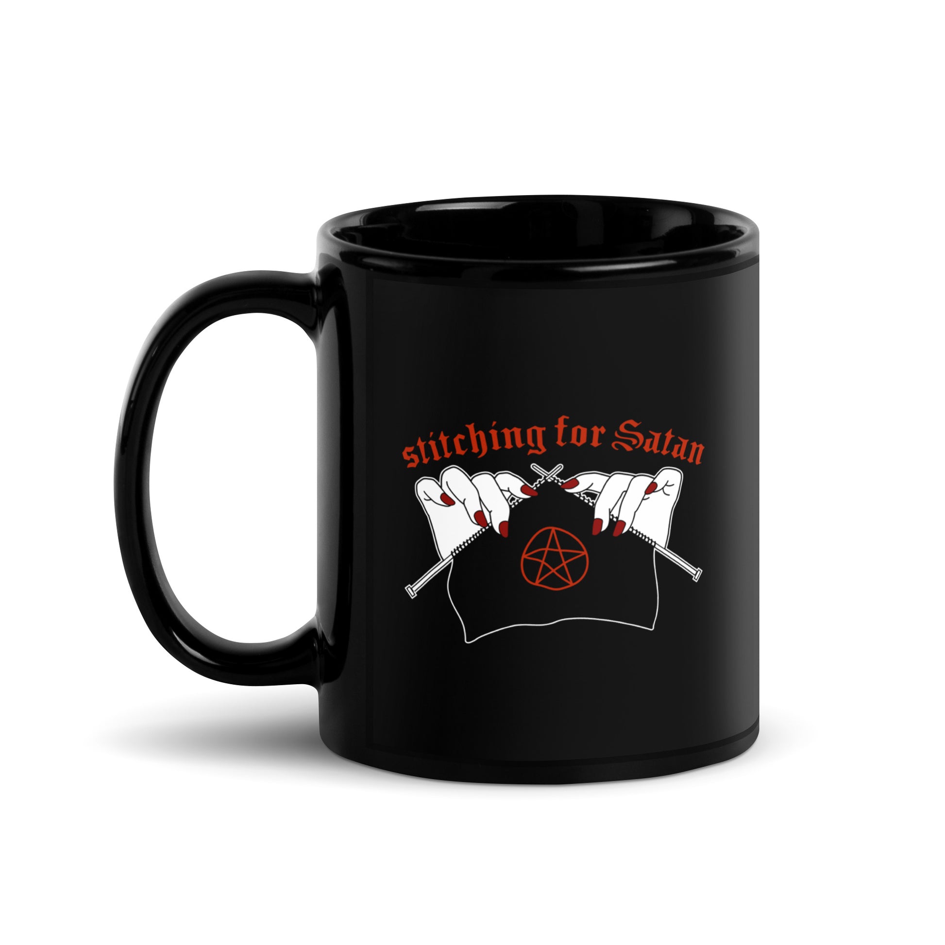 A black ceramic coffee mug featuring an illustration of pale white hands with red fingernails holding knitting needles. Fabric on the needles features a red pentagram. Text above the hands reads "stitching for Satan" in a gothc red font.