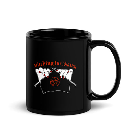 A black ceramic coffee mug featuring an illustration of pale white hands with red fingernails holding knitting needles. Fabric on the needles features a red pentagram. Text above the hands reads "stitching for Satan" in a gothc red font.
