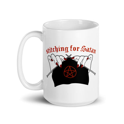 A white 15oz ceramic coffee mug featuring an illustration of pale white hands with red fingernails holding knitting needles. Fabric on the needles features a red pentagram. Text above the hands reads "stitching for Satan" in a gothc red font.