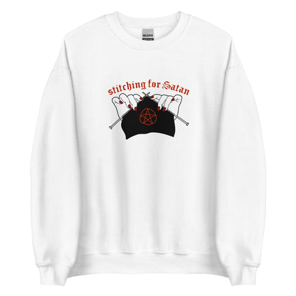 A white crewneck sweatshirt featuring an illustration of pale white hands with red fingernails holding knitting needles. Fabric on the needles features a red pentagram. Text above the hands reads "stitching for Satan" in a gothc red font.
