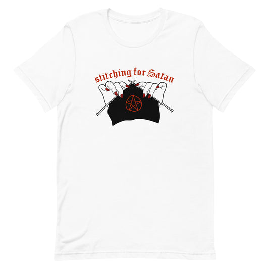 A white crewneck t-shirt featuring an illustration of pale white hands with red fingernails holding knitting needles. Fabric on the needles features a red pentagram. Text above the hands reads "stitching for Satan" in a gothc red font.