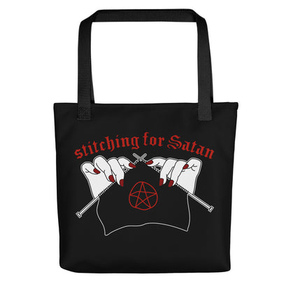 A black canvas tote bag with black handles. The bag is decorated with an illustration of pale white hands with red fingernails holding knitting needles. Fabric on the needles features a red pentagram. Text above the hands reads "stitching for Satan" in a gothc red font.