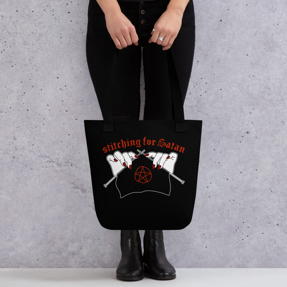 A waist-down image of a model wearing all black. The model is holding a black canvas tote bag with black handles. The bag is decorated with an illustration of pale white hands with red fingernails holding knitting needles. Fabric on the needles features a red pentagram. Text above the hands reads "stitching for Satan" in a gothc red font.