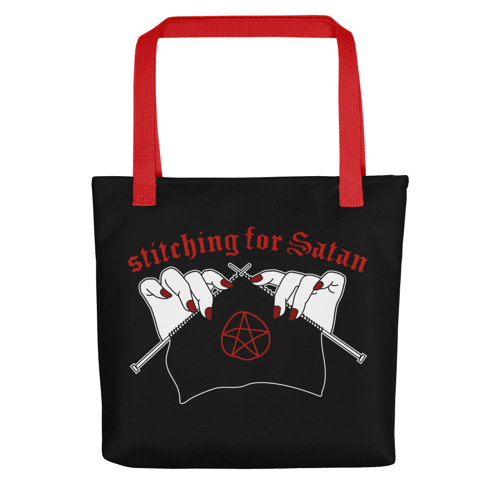 A black canvas tote bag with red handles. The bag is decorated with an illustration of pale white hands with red fingernails holding knitting needles. Fabric on the needles features a red pentagram. Text above the hands reads "stitching for Satan" in a gothc red font.