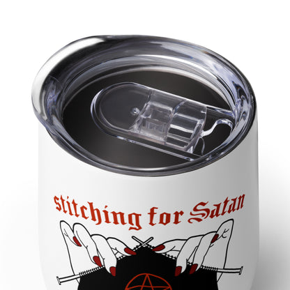 A close-up image of the lid of a white 12 ounce wine tumbler with a clear plastic lid. The tumbler is decorated with an illustration of pale white hands with painted red nails holding knitting needles. Black fabric on the needles features a red pentagram, and text above the hands reads "stitching for Satan" in a gothic red font.