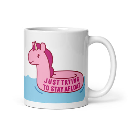 A white 11oz ceramic mug with a pink unicorn pool float floating on water and text reading "Just trying to stay afloat"
