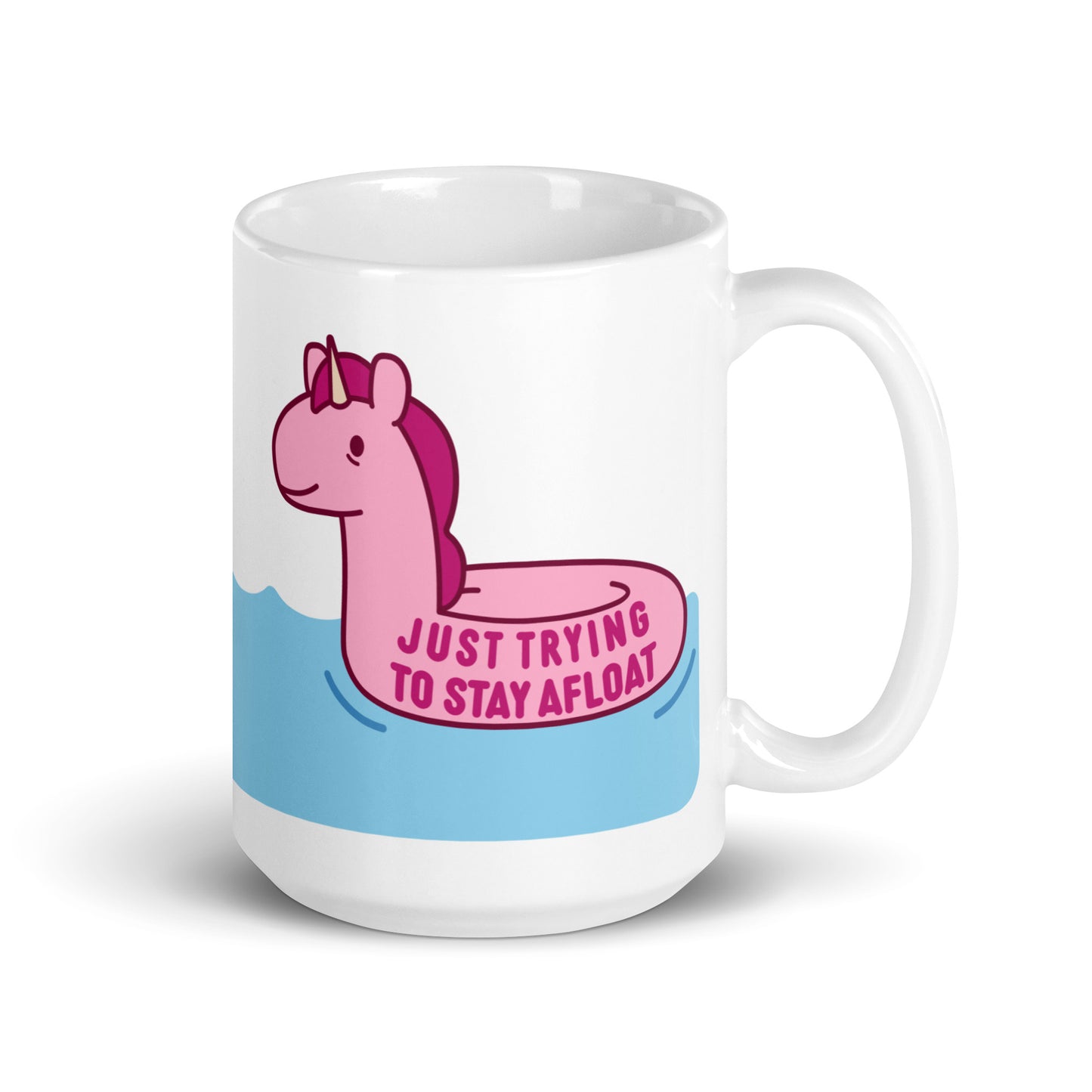 A white 15oz ceramic mug with a pink unicorn pool float floating on water and text reading "Just trying to stay afloat"