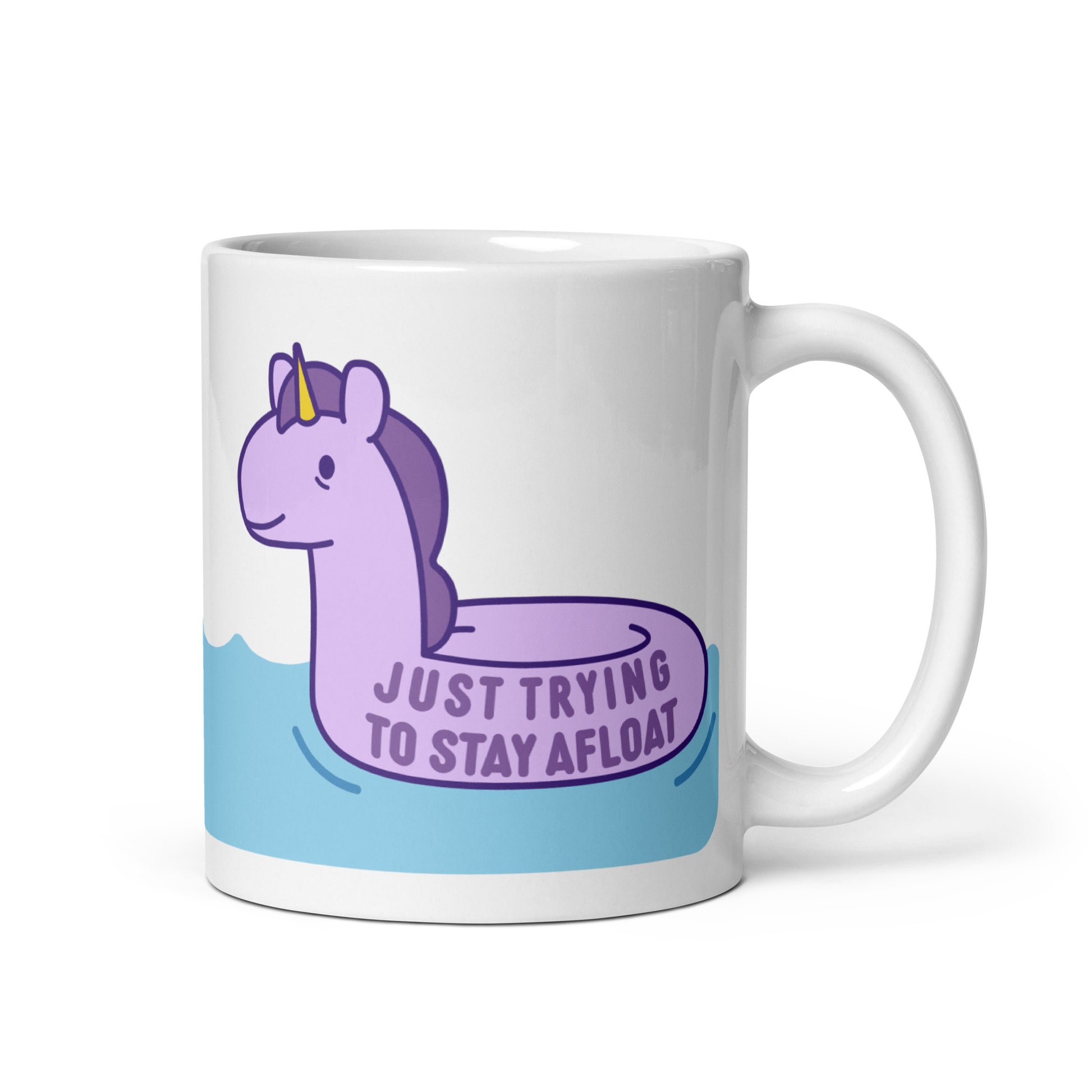 A white 11oz ceramic mug with a purple unicorn pool float floating on water and text reading "Just trying to stay afloat"