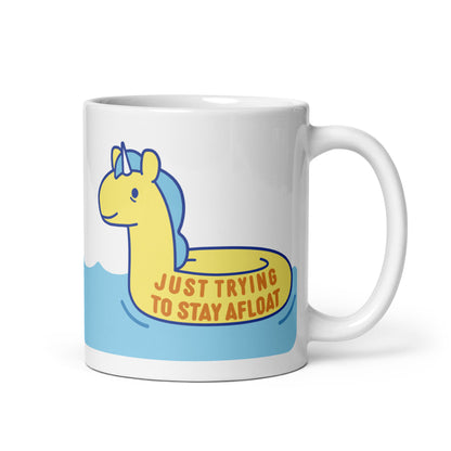 A white 11oz ceramic mug with a yellow unicorn pool float floating on water and text reading "Just trying to stay afloat"
