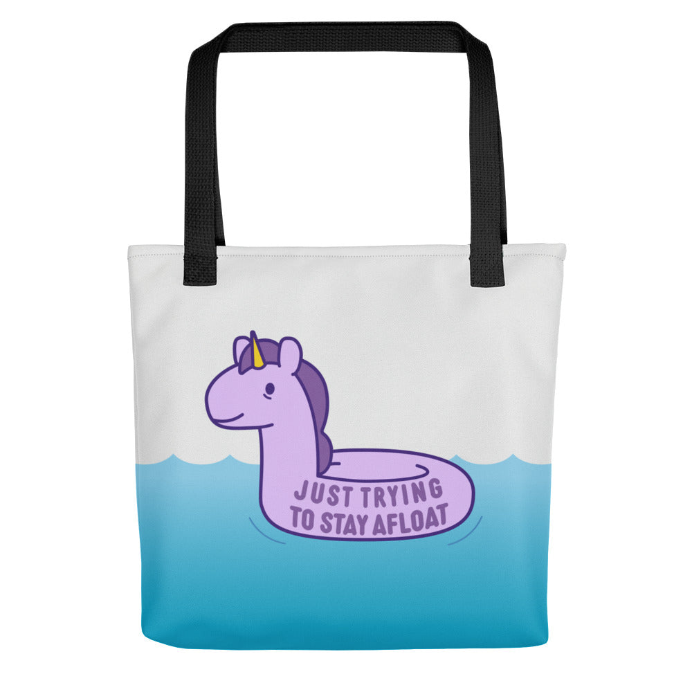 A white tote bag with black handles decorated with an image of a stressed-out unicorn floaty on water. Text on the unicorn floaty reads "Just trying to stay afloat"