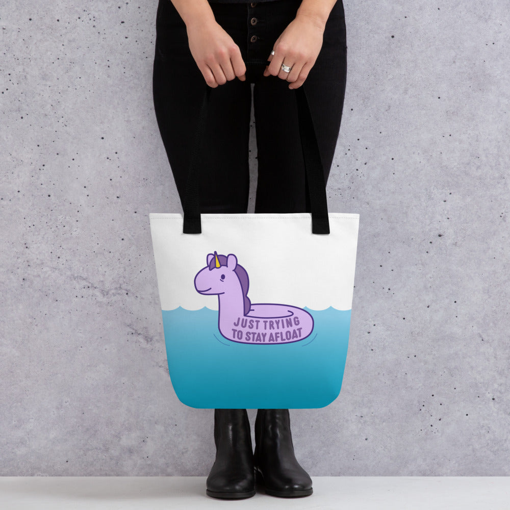 A waist-down shot of a person holding a white tote bag with black handles. The tote is decorated with an image of a stressed-out unicorn floaty on water. Text on the unicorn floaty reads "Just trying to stay afloat"