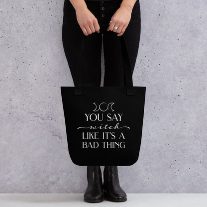 A waist-down image of a person holding a black tote bag with black handles featuring the triple goddess symbol and text reading "You say witch like it's a bad thing"