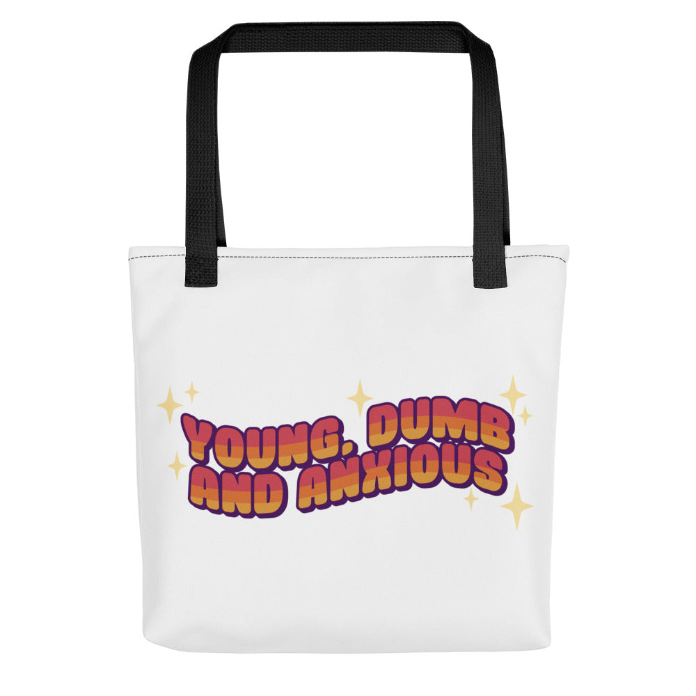 A white tote bag with black handles featuring text in a pink-to-yellow gradient, surrounded by sparkles. The text reads "Young, dumb and anxious" in a blocky font.