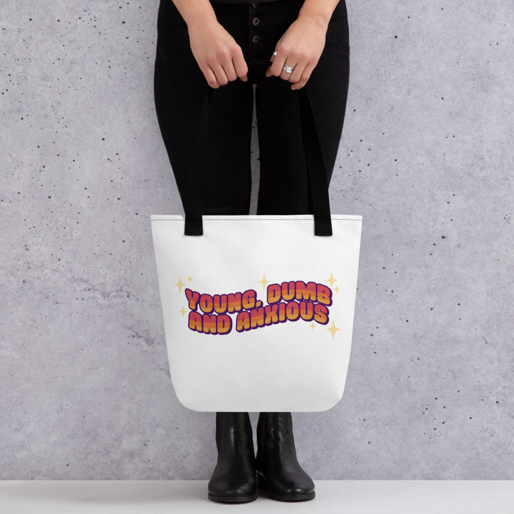 A waist-down photograph of a model holding a white totebag with black handles. The tote bag features text in a pink-to-yellow gradient, surrounded by sparkles. The text reads "Young, dumb and anxious" in a blocky font.
